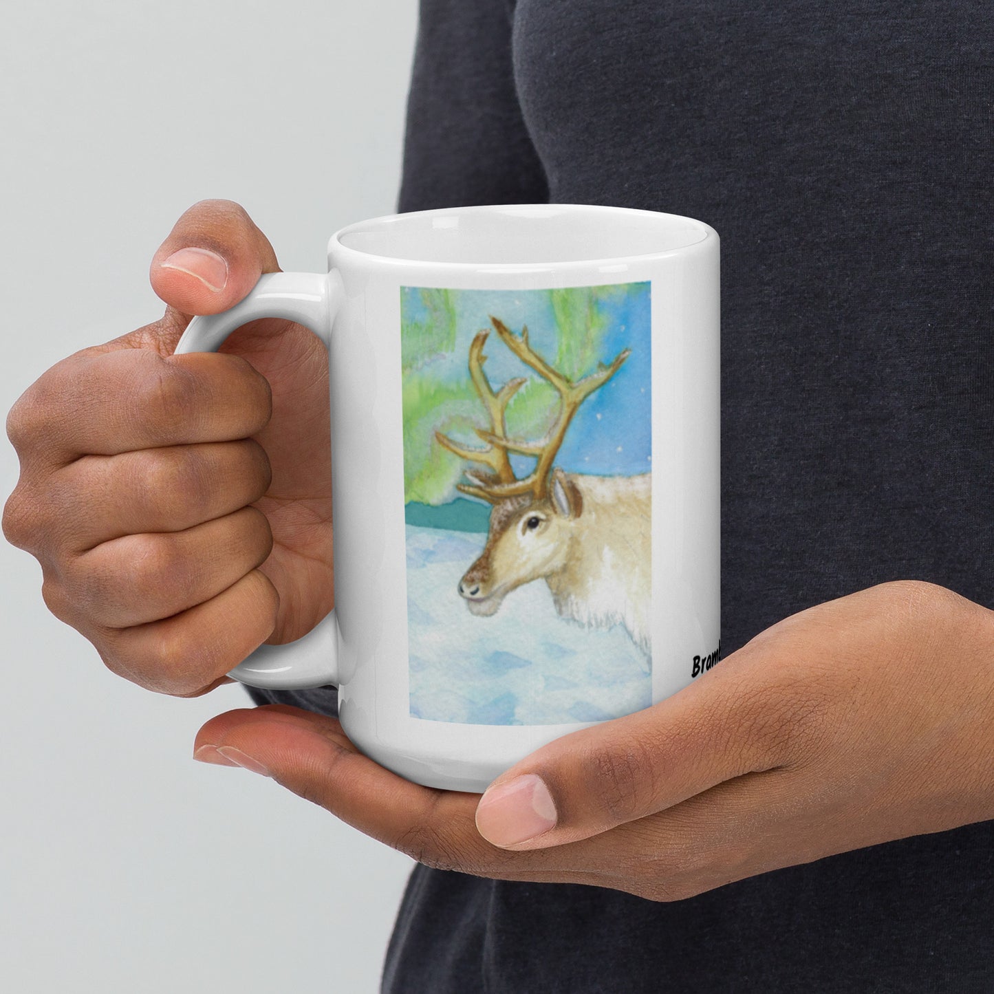 15 ounce ceramic mug featuring Northern Lights reindeer design. Double-sided print. Dishwasher and microwave safe. Shown in model's hands.