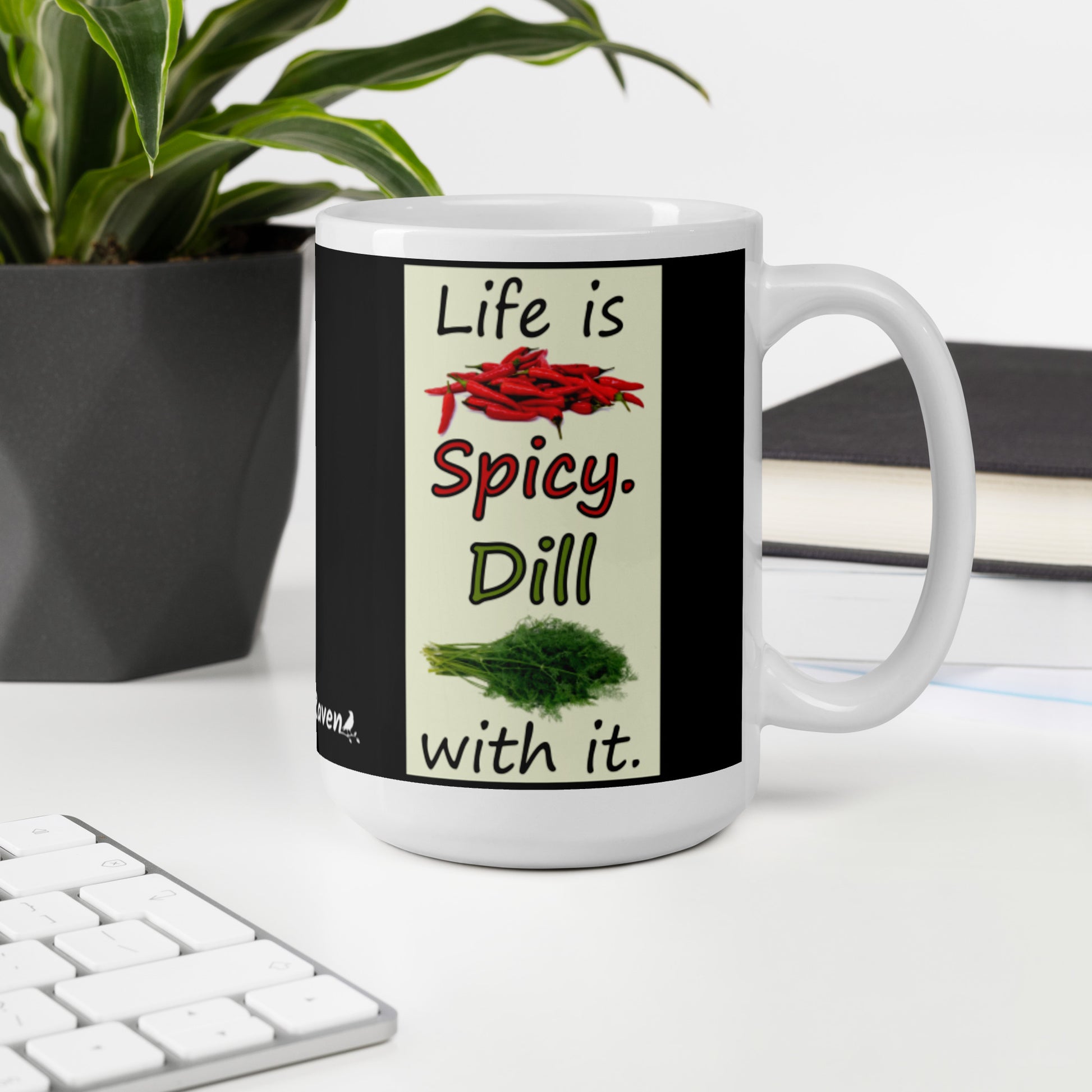 15 ounce white glossy ceramic mug. Life is Spicy. Dill with it text. Image of chili peppers and dill weed on a light yellow rectangle. Black background on mug. Shown on desktop by keyboard and potted plant.