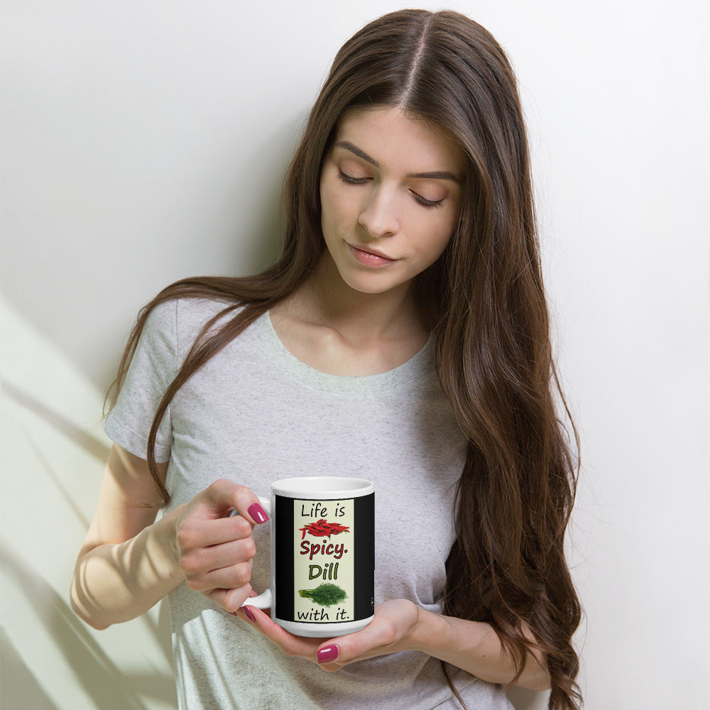 15 ounce white glossy ceramic mug. Life is Spicy. Dill with it text. Image of chili peppers and dill weed on a light yellow rectangle. Black background on mug. Shown in the hands of female model.